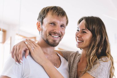 Neşeli, sakallı, beyaz tenli bir adamın, kameraya gülümseyen, neşeli bir ruh hali içinde, sevimli bir kız arkadaşının ona sarılıp mutlu, genç bir evlilikle kapalı mekanda aşk dolu duygular besleyen bir portresi.