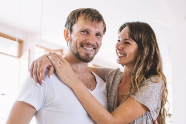 Neşeli, sakallı, beyaz tenli bir adamın, kameraya gülümseyen, neşeli bir ruh hali içinde, sevimli bir kız arkadaşının ona sarılıp mutlu, genç bir evlilikle kapalı mekanda aşk dolu duygular besleyen bir portresi.