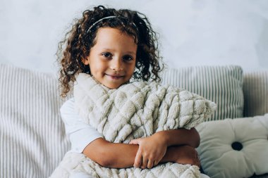 Sevimli kıvırcık kız rahat bir kanepede oturmuş, bacaklarını çaprazlamış, yumuşak yastığı kucaklıyor ve kameraya gülümsüyor.