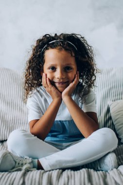 Minderleri kameraya gülümserken rahat bir koltukta otururken ellerine yaslanan kıvırcık saçlı sevimli küçük kız.