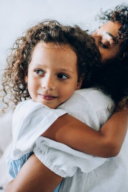Sevimli küçük kız ve annesinin yan görünüşü. İkisi de kıvırcık saçlı, kanepede oturuyor ve evde sevgiyle kucaklaşıyorlar.