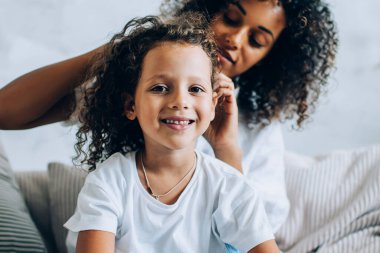 Sevimli, gülümseyen bir kız, annesiyle kanepede oturuyor kıvırcık saçlarını örüyor ve mutlu bir şekilde kameraya bakıyor evde birlikte vakit geçiriyorlar.