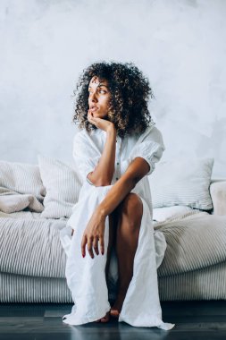 Beyaz bir evde kıvırcık saçlı, genç, şehvetli bir kadın evdeki kanepede oturmuş, gün ışığında dalgın dalgın dalgın uzaklara bakıyor.