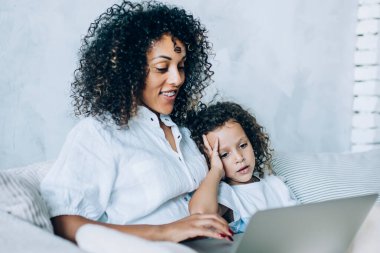 Güzel kıvırcık kadın ve tatlı kızı dizüstü bilgisayar izliyorlar. Evdeki kanepede oturuyor ve boş vakitlerinde vakit geçiriyorlar.