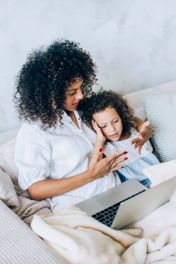 Küçük tatlı kızı olan güzel bir etnik kadın ekoseli kanepeye uzanmış dizüstü bilgisayar kullanıyor ve evde birlikte dinleniyor.