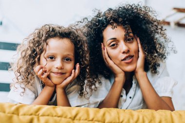 Sevimli, küçük kızı olan, yatakta uzanmış kameraya bakarken aynı kıvırcık saçlara sahip, çekici, yetişkin bir kadın.