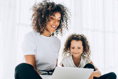 Güzel, kıvırcık bir kadın ve gülümseyen bir kız, yumuşak yastıklarda oturup dizüstü bilgisayar izliyorlar. Evde boş vakitlerinde vakit geçiriyorlar.