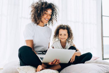 Güzel, kıvırcık bir kadın ve gülümseyen bir kız, yumuşak yastıklarda oturup dizüstü bilgisayar izliyorlar. Evde boş vakitlerinde vakit geçiriyorlar.