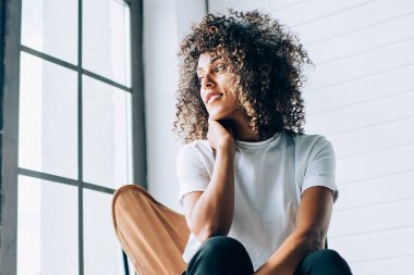 Pencere kenarındaki rahat koltukta oturan ve yumuşak ışıkta dalgın dalgın bakan şehvetli etnik kadın.