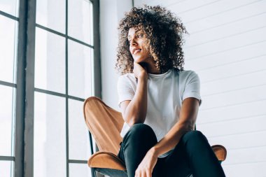 Pencere kenarındaki rahat koltukta oturan ve yumuşak ışıkta dalgın dalgın bakan şehvetli etnik kadın.