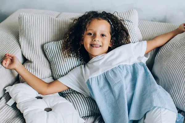 Yukardan gülümseyen küçük tatlı bir kız, büyük yumuşak bir kanepede uzanıyor ve mutlu bir şekilde kameraya bakıyor.
