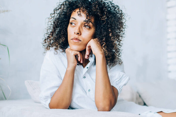 Young ethnic woman with curls lounging on comfy couch at home leaning on hands and looking dreamily away in soft light