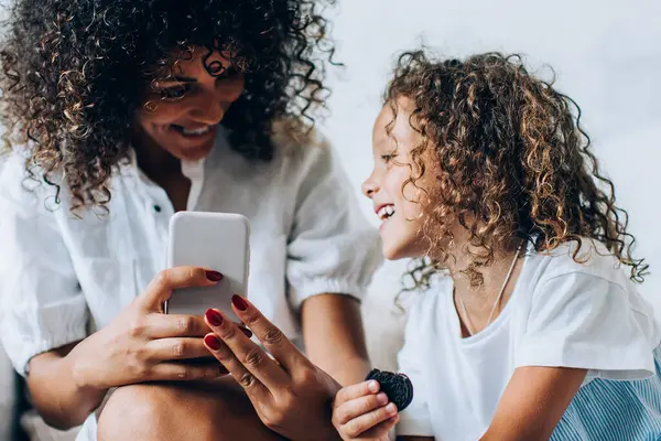 Evdeki rahat kanepede uzanıp birlikte vakit geçirirken akıllı telefon kullanan güzel, etnik bir kadın ve tatlı küçük bir kız.