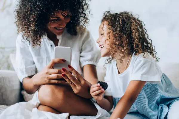 Evdeki rahat kanepede uzanıp birlikte vakit geçirirken akıllı telefon kullanan güzel, etnik bir kadın ve tatlı küçük bir kız.