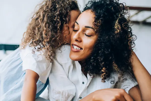 Güzel etnik kıvırcık kadın mutlu kızını okşuyor rahat bir yatakta sarılıyor ve gözlerini kapalı tutuyor evde vakit geçiriyor.