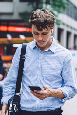 Finansal web sitesinde video izleyen beyaz erkek tüccar modern hücresel cihazda 4G kablosuz bağlantıyla bağlandı, elektronik kulaklıklı deneyimli bir işadamı ofise doğru yürüyordu.