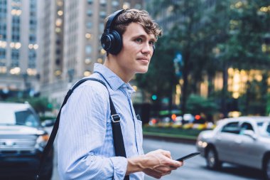 Resmi olarak başarılı bir erkek girişimci modern akıllı telefon cihazını elinde tutuyor ve müzik uygulamasını ses yayını için kulaklıklar aracılığıyla ses yalıtımı için kullanıyor.