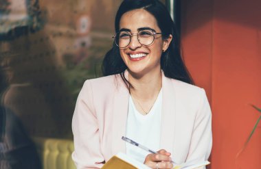 Gözlüklü mutlu yeniyetme kız göz korumasını sağladığı için gülümserken üniversite kompozisyonu hazırlıyor. Heyecanlı bir fikirle şehir merkezindeki kahve dükkanının yanında oturuyor. Notnotlu pozitif öğrenci.