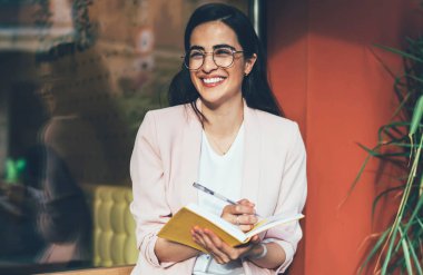 Gözlüklü mutlu yeniyetme kız göz korumasını sağladığı için gülümserken üniversite kompozisyonu hazırlıyor. Heyecanlı bir fikirle şehir merkezindeki kahve dükkanının yanında oturuyor. Notnotlu pozitif öğrenci.