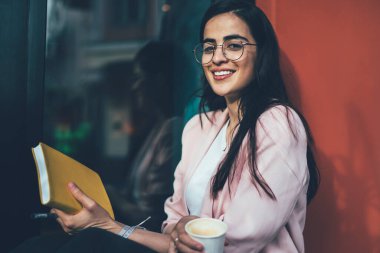 Neşeli İspanyol erasmus öğrencilerinin portresi kahveyle dinleniyor eğitim kitapçığıyla planlama sürecinin peşinden gitmek için dışarıda kameraya gülümseyen bilgi notları olan pozitif kadın.