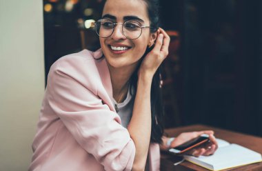 Şık klasik gözlüklü mutlu bayan öğrenci sokak kafeteryasında okuduğu için kahkahalar atıyor. Ders çalışması için cep telefonu kullanan gözlüklü pozitif Gürcü kadın.