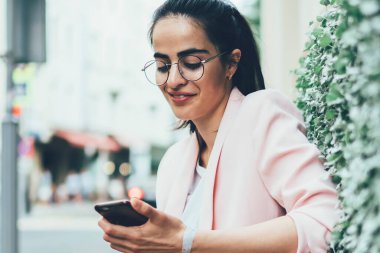 Mutlu blogcu akıllı telefon cihazıyla ödemeyi 4G internet üzerinden yapıyor açık hava bankında dinleniyor, şık ceketli ve gözlüklü başarılı genç kadın cep telefonuyla arkadaşça sohbet ediyor.