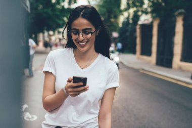 Sosyal ağlarda profilini güncelleyen olumlu bayan öğrenci şehir merkezinde üniversite arkadaşını beklerken, mutlu İspanyol kadın cep telefonu uygulaması üzerinden sohbet etmekten hoşlanıyor