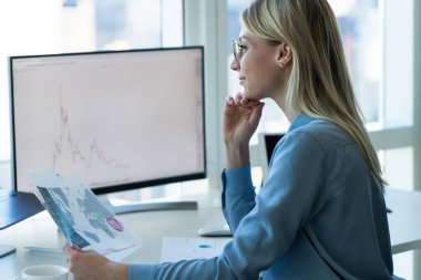 Elinde kağıt harita olan gülümseyen sarışın iş kadınının yan görünüşü ve ofiste çalışırken dizüstü bilgisayarına bakışı.