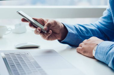 Mavi tişörtlü, cep telefonlu bir adamın çalışma masasında modern bir bilgisayar ve dizüstü bilgisayarla oturması.