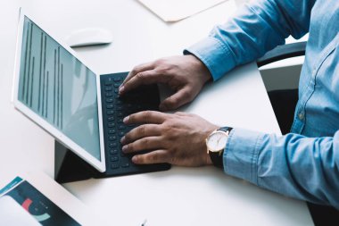 Mavi gömlekli bir yetişkinin beyaz bilgisayar masasında oturup tabletle taşınabilir klavyeye yazışının kırpma kenarı görüntüsü
