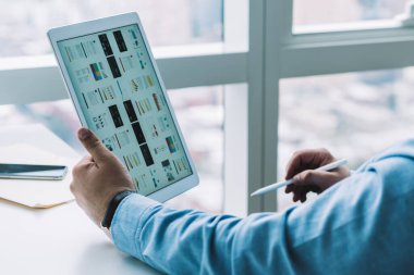 Ofisteki bilgisayar masasında oturan ve internet üzerinde çalışırken tablet izleyen mavi gömlekli adam yan görünümü