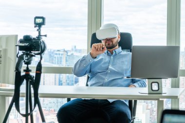 VR gözlüklü yetişkin adam modern ofiste bilgisayar masasında oturuyor ve video kamerayla sanal gerçeklikte çalışıyor.