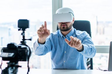 Modern profesyonel bir adam bilgisayar ve dizüstü bilgisayarla masada otururken kameraya işaret ederken sanal gerçeklik gözlüğü takıyor.