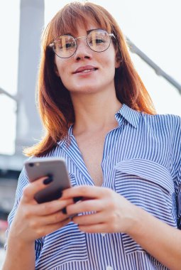 Elinde cep telefonu olan beyaz kadın gezginin yarım boy portresi kameraya bakarken web sitesinde konum bilgisi, milenyum zaman ve turizm kavramına göz atıyor.