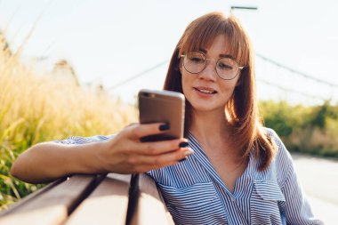 Kullanışlı bilgilerle birlikte web sayfasında klasik gözlük paylaşımı yapan beyaz blogcu, e-postaları kontrol eden ve akıllı telefon aygıtıyla mesaj gönderen çekici kadın şehir bankında oturuyor.