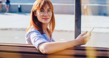 Gözlüklü çekici bir kadın bankta oturuyor, akıllı telefondan sosyal ağlarda sohbet ediyor, güneşli bir günde açık havada oturup cep telefonu uygulamasında postaları kontrol eden kadın blogcunun portresi.