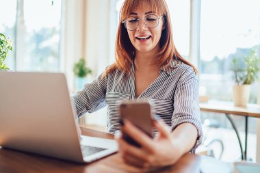 Klasik gözlüklü mutlu hipster blogcu medya dosyalarını paylaşmak için hızlı kurulum mobil uygulamasından iyi hissediyor, Kafkasyalı kızıl saçlı kadın cep telefonu aygıtı bildirimini kontrol ediyor