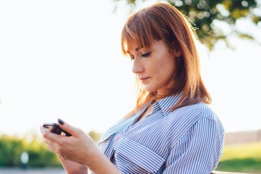 Bin yıllık düşünceli kızıl saçlı kadın parkta bankta oturan cep telefonuyla postaları kontrol etmeye odaklanmış. Kafkasyalı genç hippi kız, bankacılık ve online ödemeler için akıllı telefon uygulaması kullanıyor.