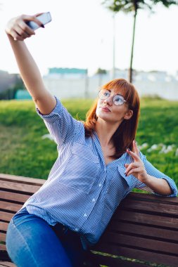 Açık havada akıllı telefon kamerasıyla selfie çeken çekici genç bir kadın, göz giysili kızıl saçlı kadın cep telefonu uygulaması ile blogu için fotoğraf çekiyor.