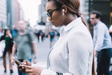 Kalabalık şehir sokaklarının bulanık arka planında yürürken şık giyinmiş, gözlüklü, müzik dinleyen ve akıllı telefon kullanan ciddi Afrikalı Amerikalı kadınların yan görüntüsü.