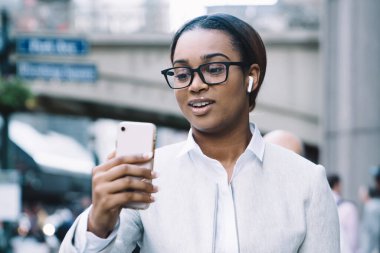 Beyaz takım elbiseli, kablosuz kulaklıklı, sokakta yürürken cep telefonuyla konuşan akıllı, Afro-Amerikalı bir kadın.