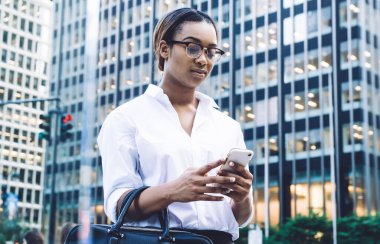 Siyah çantalı, gözlüklü, şık elbiseli, beyaz perdeye odaklanmış ve caddede bulanık şehir hayatına karşı dururken akıllı telefon kullanan düşünceli Afro-Amerikan kendinden emin bir kadından.