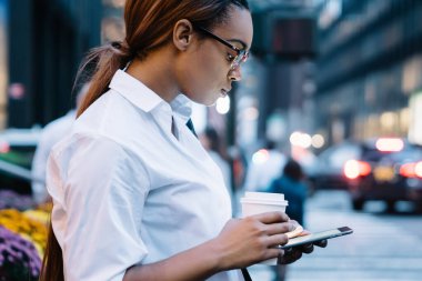 Odaklanmış Afrika kökenli Amerikalı bir kadın elinde bir bardak içecekle şehir caddesinde yol kenarında dururken modern tabletlere göz atmaya gidiyor.