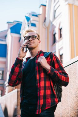 Entellektüel gülümseyen kızıl saçlı erkek işaretli kırmızı ve siyah gömlekli cep telefonuyla konuşuyor şehirdeki granit barajın üzerinde duruyor.