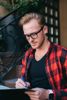 Odaklanmış kızıl saçlı erkek işaretli kırmızı ve siyah gömlekli. Not defterine not alıyor. Yazın şehirde granit merdivenlerde oturuyor.