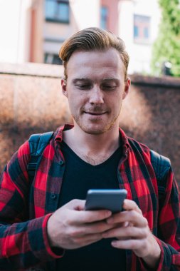 Kızıl sakallı genç erkek işaretli kırmızı ve siyah gömlekli. Yazın şehirde gezintiye çıkan cep telefonuyla sörf yapıyor.