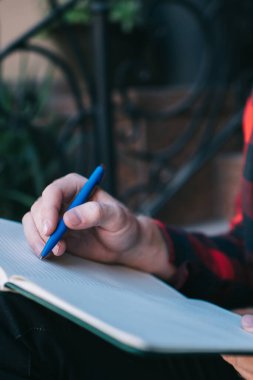 Kırmızı ve siyah tişörtlü bir adam yazın şehirde merdivenlerde oturan kalemle not defteri tutuyor.