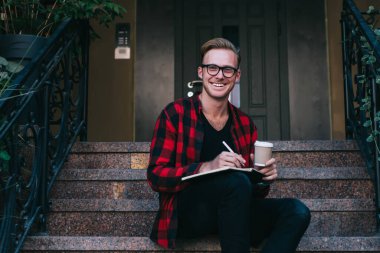 Neşeli, güler yüzlü, kızıl saçlı, işaretlenmiş kırmızı ve siyah tişörtlü bir adam. Yazın şehirde granit merdivenlerde oturan kalemle not alıyor.