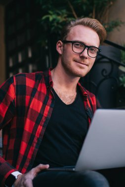 Günlük kıyafetleri ve gözlükleri olan yakışıklı bir adam dizüstü bilgisayar kullanıyor. Yeni başlayanlar için evde oturup kameraya bakıyor.