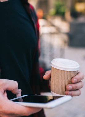 Günlük kıyafetlerle gezen, sosyal medyayı izleyen cep telefonu kullanan, şehrin dışında gezen, sıcak kahve içen genç adamın görüntüsü.